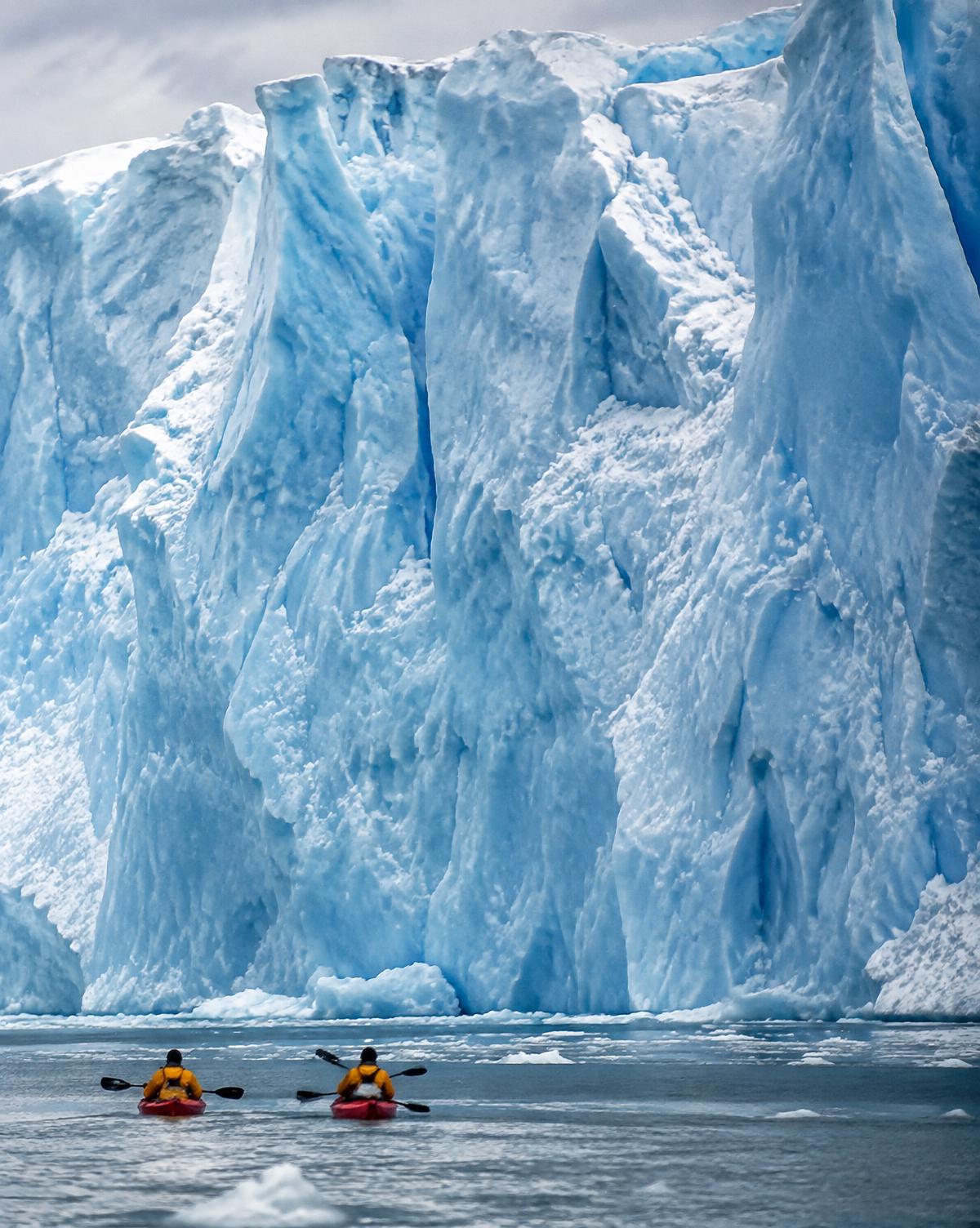 Two kayakers paddling beside an immense glacier wall